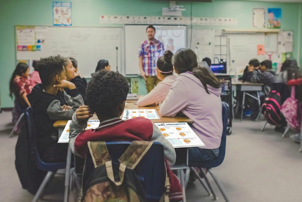 A real primary school classroom with a teacher at the board and Classprint worksheets on the students' desks